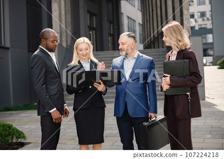 Confident businesspersons standing in front of modern office building. Men and women talking and holding documents. Banking and financial market concept. 107224218