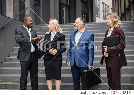 Confident businesspersons standing in front of modern office building. Men and women talking and holding documents. Banking and financial market concept. 107224219