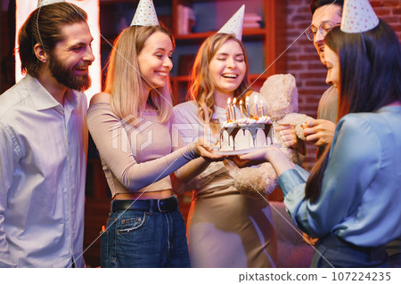 Young friends standing in indoors in evening party red light. Everybody wearing party cones. Boys and girls holding a birthday cake with a candles. 107224235