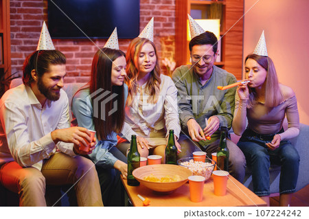 Young friends sitting on a coach in evening party red light. Everybody wearing party cones. Friends holding a paper cups with beverages and eating popcorn. 107224242