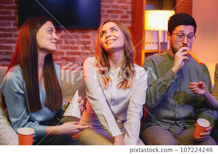 Young friends sitting on a coach in evening party red light. Blonde girl in a middle has a birthday. Friends, party and celebration concept. 107224246