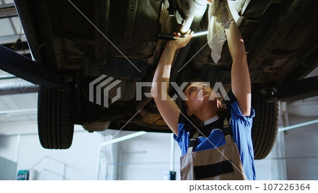 Certified technician working on suspended car in garage, checking components during routine maintenance. Auto repair shop worker walking underneath vehicle, examining it using work light Certified technician working on suspended car in garage, checking components during routine maintenance. Auto repair shop worker walking underneath vehicle, examining it using work light 107226364