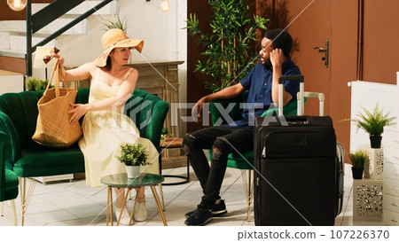 Couple sitting in lounge area lobby at hotel, waiting to do registration and check in process. Guests on honeymoon summer holiday arriving at tropical seaside resort, waiting for concierge services. Couple sitting in lounge area lobby at hotel, waiting to do registration and check in process. Guests on honeymoon summer holiday arriving at tropical seaside resort, waiting for concierge services. 107226370