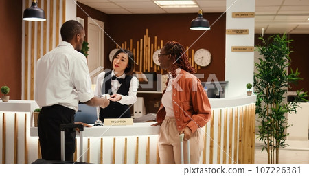 Friendly bellhop employee showing tourists the way to their room while carrying luggage after check in process is over. Stressed hotel manager verifying guests booking paperwork 107226381