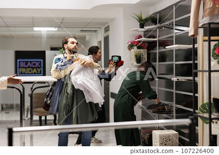 Stressed Black Friday obsessed diverse people running with clothes around fashion store with empty shelves, customers hunting for bargains during big seasonal sales in shopping mall 107226400