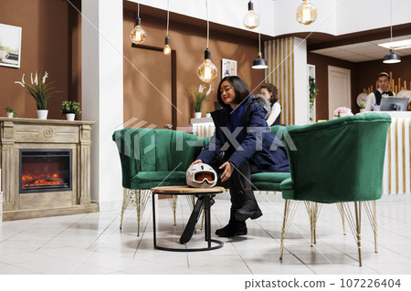 Young female tourist sitting on sofa in lounge area of luxurious winter resort waiting for accommodation. Enthusiastic woman placing ski helmet on table of hotel lobby ready for wintertime activities. Young female tourist sitting on sofa in lounge area of luxurious winter resort waiting for accommodation. Enthusiastic woman placing ski helmet on table of hotel lobby ready for wintertime activities. 107226404