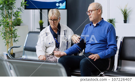 Senior medic measuring patient hypertension using tonometer instrument, checking hypotension during healthcare appointment in hospital waiting room. Elderly man having cardiac exam 107226407