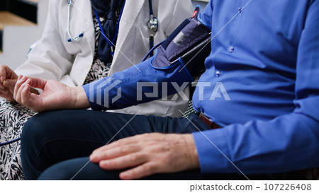 Elderly medic using medical tonometer to measure patient pulse pressure and hypertension, doing cardiology examination in hospital waiting room. Senior man attending checkup visit. Medicine concept Elderly medic using medical tonometer to measure patient pulse pressure and hypertension, doing cardiology examination in hospital waiting room. Senior man attending checkup visit. Medicine concept 107226408