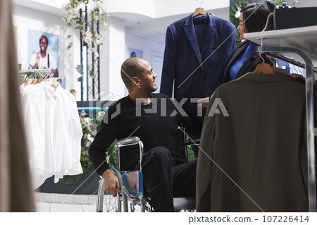 Smiling arab man buyer with disability examining trendy jacket style while seller holding hanger with apparel in clothing store. Boutique employee and client in wheelchair choosing outfit together 107226414