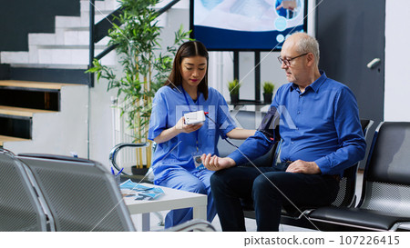 Nurse checking patient cardiac pulse during cardiology examination in hospital waiting area. Asian assistant measuring senior man blood pressure and hypertension using tonometer instrument 107226415