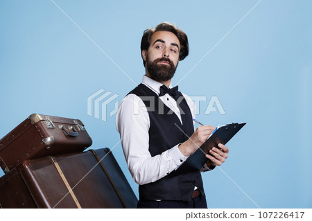 Classy bellhop creating guestlist at hotel, using archive record files on clipboard to keep evidence. Stylish doorkeeper taking notes with information on papers, verifying bulletpoints. 107226417