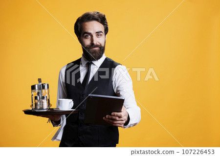 Personnel carries food bill on tablet, serving customers with cup of coffee at luxury restaurant in studio. Restaurant staff gives tab to clients on modern device, posing against yellow background. 107226453