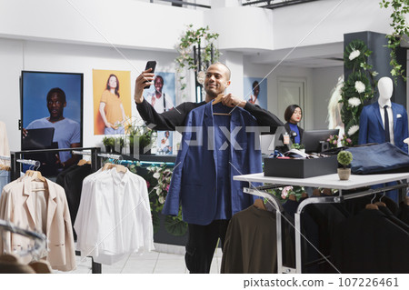 Young arab man internet influencer live streaming using smartphone in clothing store while showcasing jacket. Boutique client choosing outfit and showing apparel on hanger on mobile phone front camera Young arab man internet influencer live streaming using smartphone in clothing store while showcasing jacket. Boutique client choosing outfit and showing apparel on hanger on mobile phone front camera 107226461