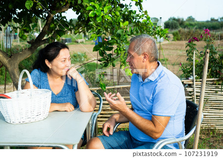 Mature couple sitting at table in backyard garden and talking Mature couple sitting at table in backyard garden and talking 107231919