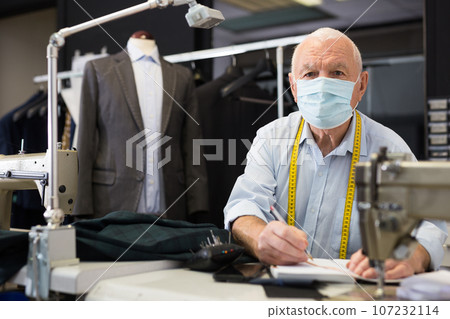 Portrait of an elderly tailor in protective mask making notes in notebook 107232114
