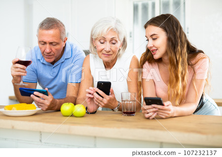 Elderly parents and adult daughter surf the internet using mobile phones in kitchen Elderly parents and adult daughter surf the internet using mobile phones in kitchen 107232137