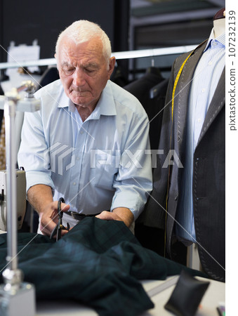 Elderly tailor cuts fabric with scissors at workplace in workshop Elderly tailor cuts fabric with scissors at workplace in workshop 107232139