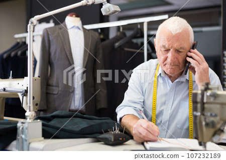 Mature tailor wearing protective mask talking on a mobile phone and taking notes in notebook 107232189