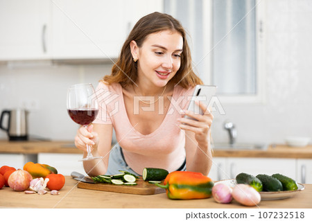 Cheerful woman chatting with friends on smartphone and drinking wine while standing near table in cozy home kitchen 107232518