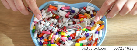 Women's hands hold knife and fork over heart-shaped plate with bunch of different pills. Women's hands hold knife and fork over heart-shaped plate with bunch of different pills. 107234635