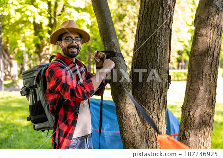 Smiling dark-haired bearded young traveler standing near the tree Smiling dark-haired bearded young traveler standing near the tree 107236925