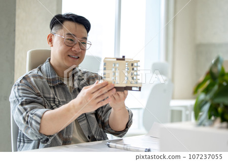 Man with house model printed with 3D printer in light office of creative lab. Man with house model printed with 3D printer in light office of creative lab. 107237055