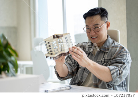Man with house model printed with 3D printer in light office of creative lab. Man with house model printed with 3D printer in light office of creative lab. 107237057