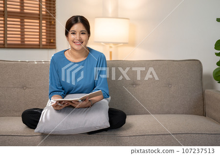 A beautiful Asian woman in casual clothes sits on a couch with a book in her hand. A beautiful Asian woman in casual clothes sits on a couch with a book in her hand. 107237513