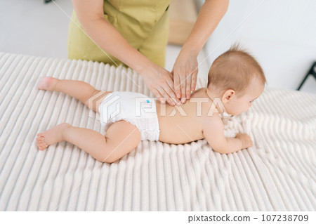 High-angle view of loving mother hands massaging little baby. Close up cropped shot of unrecognizable masseuse female hands doing massage to newborn. Back view of cute infant baby lying on stomach. 107238709