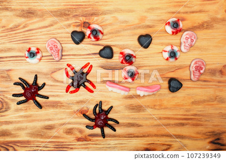 Halloween jelly candies in skulls, vampire teeth, black heart, bones, spiders, on a wooden background with a hard shadow. Festive pattern of sweets, treats. 107239349