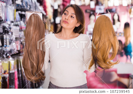 Positive young woman choosing natural wig at specialty boutique Positive young woman choosing natural wig at specialty boutique 107239545