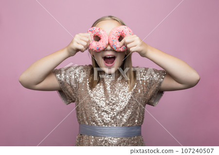 Happy child girl holding two tasty pink donuts on bright pink background, fashion portrait.  107240507