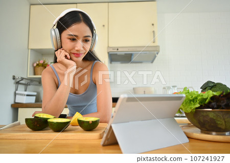 Charming young woman following online recipe on digital tablet while preparing healthy, vegetarian food in kitchen at home 107241927