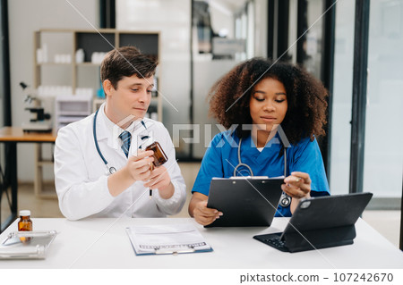 Focused mature male doctor and African female nurse look at tablet and laptop discuss anamnesis together. Concentrated diverse medical professionals in hospital. 107242670