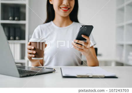 Beautiful asian woman holding and using digital tablet while drinking coffee at home office Beautiful asian woman holding and using digital tablet while drinking coffee at home office 107243703