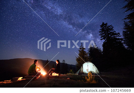 Nocturnal camping excursion in mountains, basking under twinkling stars. Man and woman, adventurous travelers, find repose by the bonfire and their illuminated tent nestled amidst the woods. 107245669