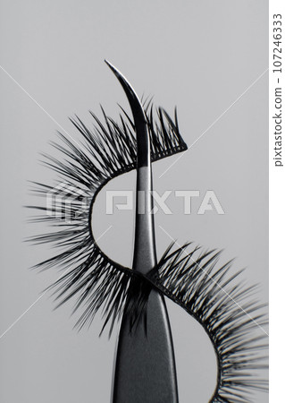 Makeup artist tools and accessories. Close-up image of metal tweezers with eyelashes for extension on gray background. Makeup artist tools and accessories. Close-up image of metal tweezers with eyelashes for extension on gray background. 107246333