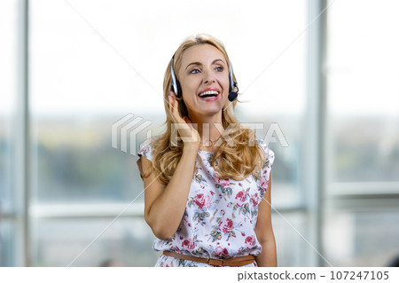 Portrait of smiling blond woman wearing headset indoors. Blurred window in the background. 107247105