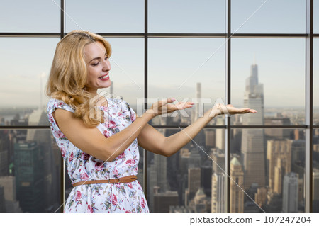 Sexy caucasian blonde woman shows copy place on her palm standing indoors. Checkered window with cityscape view in the background. Sexy caucasian blonde woman shows copy place on her palm standing indoors. Checkered window with cityscape view in the background. 107247204