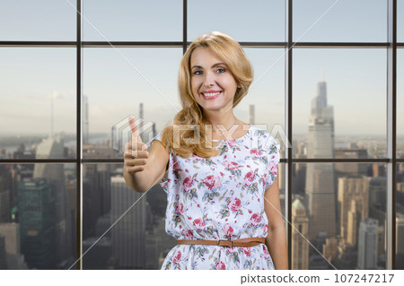 Happy blonde middle aged woman shows thumb up. Checkered window with cityscape view. Happy blonde middle aged woman shows thumb up. Checkered window with cityscape view. 107247217