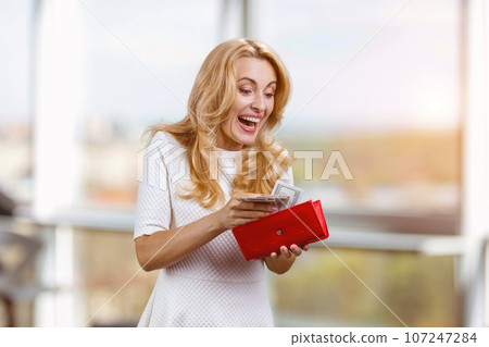 Happy excited blonde caucasian woman withdraws cash money from the red wallet. Blurred indoor window in the background. 107247284