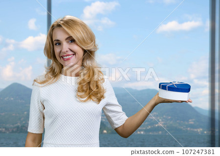 Happy middle aged blonde woman holds a blue gift box in one hand. Window with mountain scape in the background. 107247381