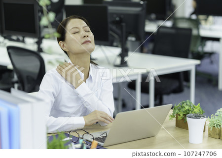 A middle-aged woman holding her shoulders in the office with stiff shoulders 107247706