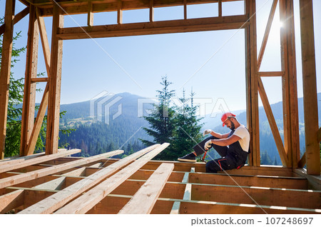 Carpenter constructing wooden frame two-story house near the forest. Bearded man holding hammer, dressed in protective helmet and overalls. Concept of ecological modern construction. Carpenter constructing wooden frame two-story house near the forest. Bearded man holding hammer, dressed in protective helmet and overalls. Concept of ecological modern construction. 107248697