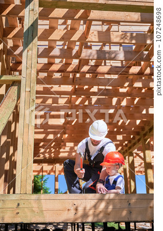 Father with toddler son building wooden frame house. Man instructing his son on the art of hammering nails on construction site, wearing helmets and overalls on sunny day. Carpentry and family concept Father with toddler son building wooden frame house. Man instructing his son on the art of hammering nails on construction site, wearing helmets and overalls on sunny day. Carpentry and family concept 107248698