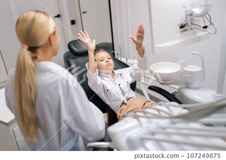 Medium shot of stressed little girl capricious and fooling sitting on dental chair and having dental treatment in dentistry clinic. Concept of children teeth treatment, pediatric checkup. Medium shot of stressed little girl capricious and fooling sitting on dental chair and having dental treatment in dentistry clinic. Concept of children teeth treatment, pediatric checkup. 107249875