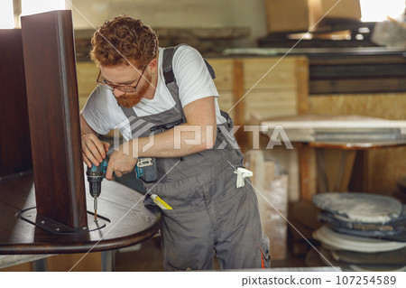 Professional carpenter fixing table with electric drill standing in carpentry workshop  107254589