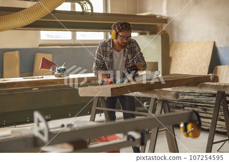 Professional carpenter in glasses is cutting wood with chainsaw on carpentry workshop 107254595