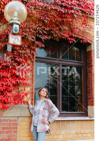 a beautiful girl stands against the background of the window of an old European house, entwined with a floating red color in autumn. Tourism & Travel Concept. Nice portrait of a young woman, in boho 107254626