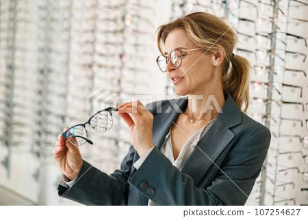 Woman in jacket chooses between two glasses in an optician's shop Woman in jacket chooses between two glasses in an optician's shop 107254627